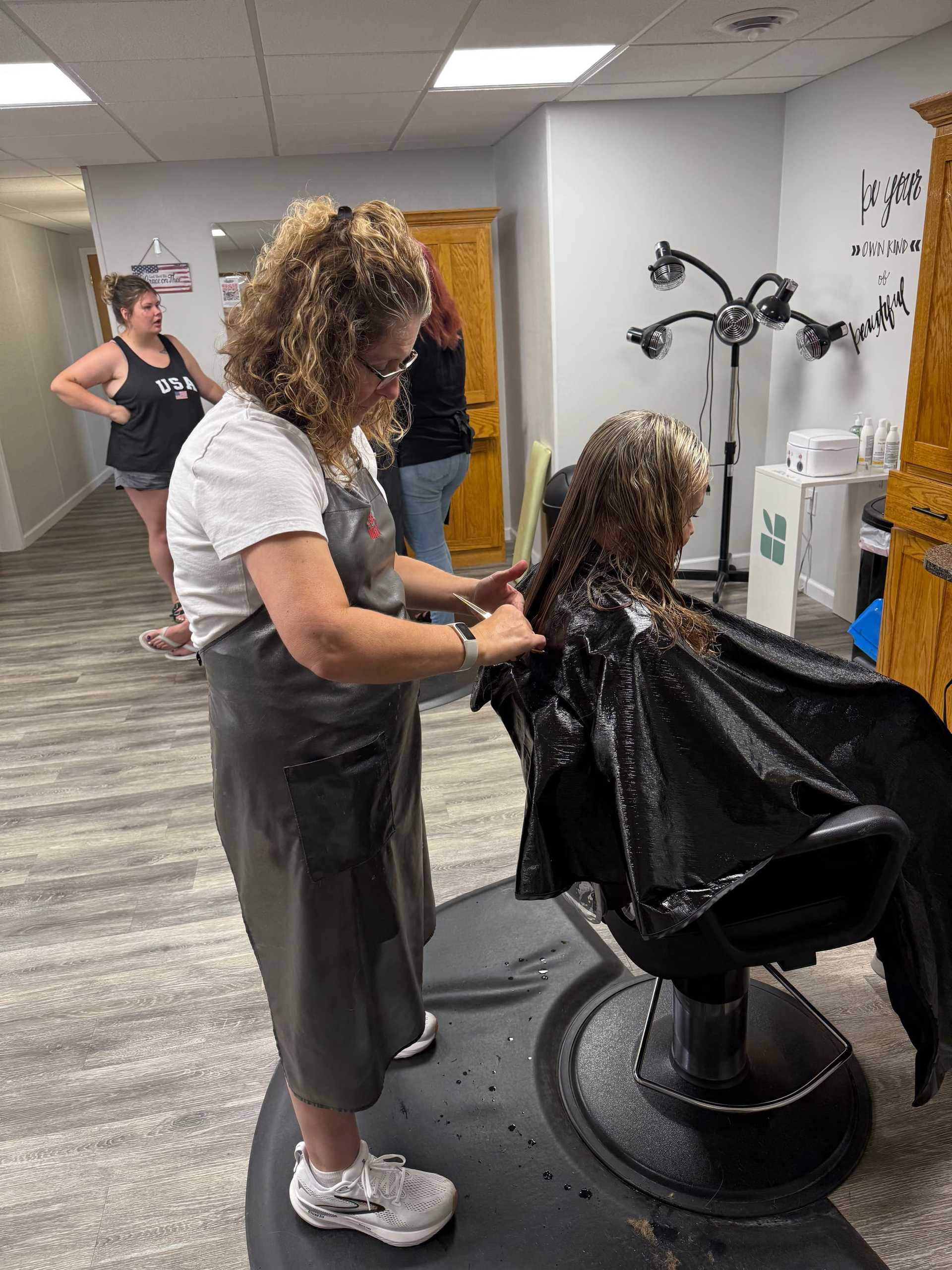 Hairdresser styling a client's wet hair in a salon, with others waiting nearby.