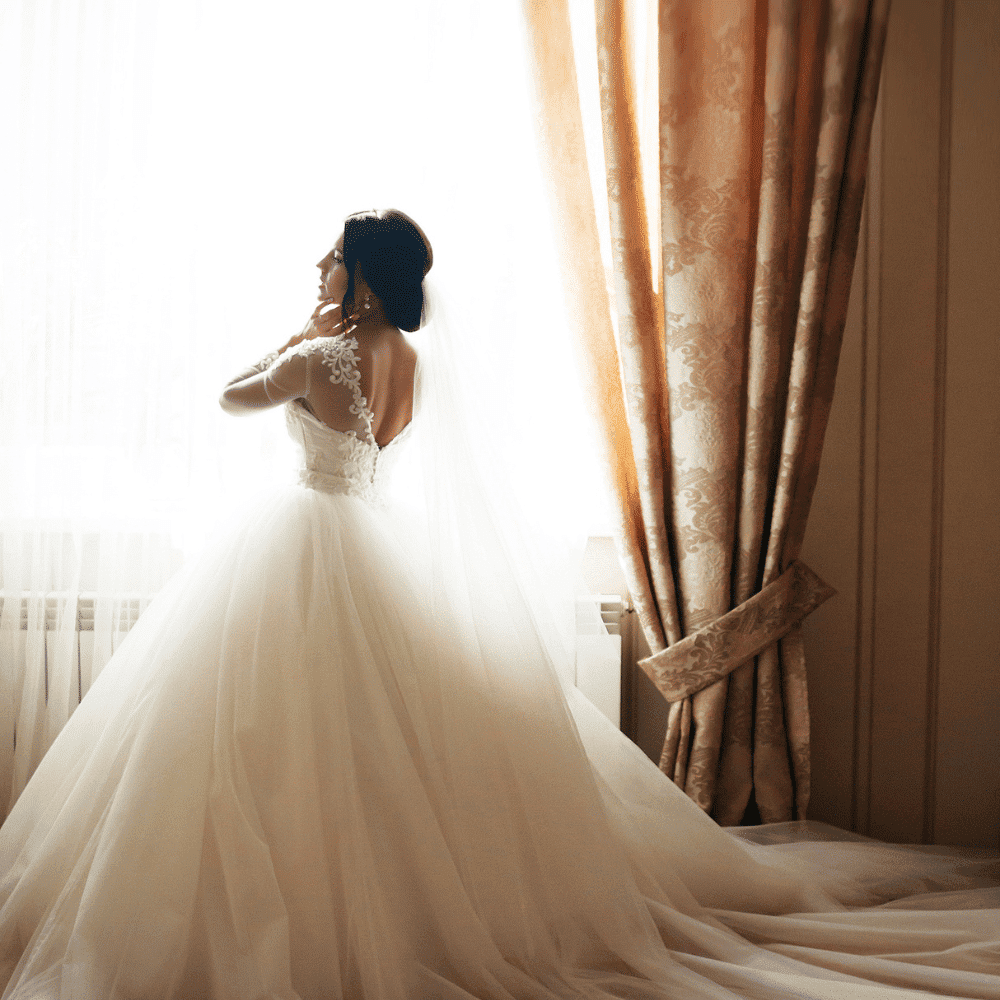 Bride in elegant white gown standing by sunlit window, looking outside.